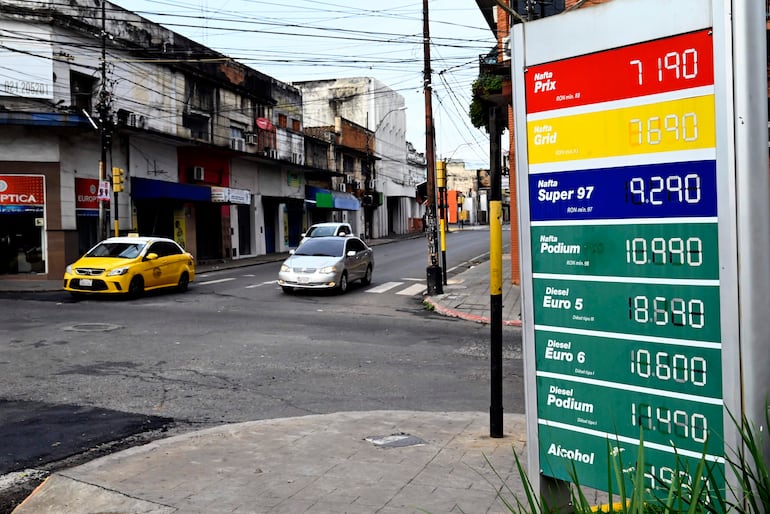 Cartel iluminado en Asunción con precios de combustibles, mientras un taxi amarillo y un automóvil blanco cruzan la intersección.