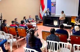 Hombre hablando con micrófono mientras 15 personas lo escuchan atentamente en un salón con banderas de Paraguay de fondo.