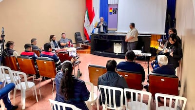 Hombre hablando con micrófono mientras 15 personas lo escuchan atentamente en un salón con banderas de Paraguay de fondo.