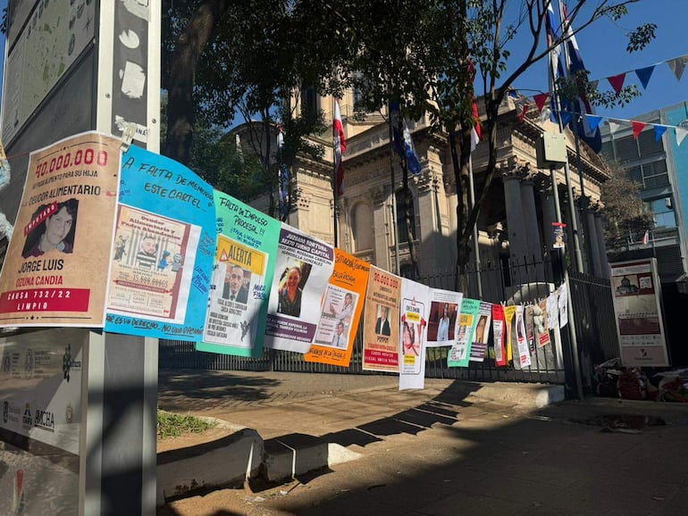 Carteles colgados en reja, sin personas visibles. Edificio destacado al fondo, ambiente tranquilo y soleado.