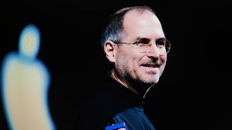 Steve Jobs sonriente, vestido con su habitual suéter negro, sostiene un nuevo iPhone mientras el público aplaude emocionado.