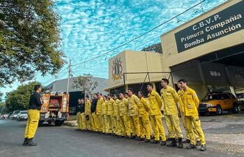 Grupo de bomberos uniformados en amarillo, escuchando a un oficial en uniforme negro en el cuartel K1, con camión de bomberos al fondo.