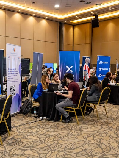 Mujer de chaqueta azul y hombre de camiseta burdeos conversan en feria de empleo, rodeados de banners de Manpower y Kemsa.