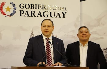 Adolfo Vallejos en traje oscuro habla al micrófono, mientras otro hombre escucha atento en conferencia del Gobierno del Paraguay.