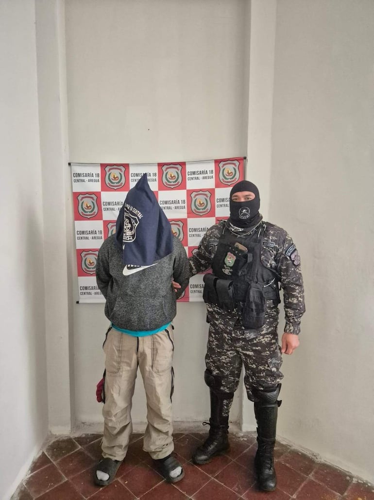 Hombre con paño oscuro siendo sostenido por un militar en uniforme en la comisaría 18 de Areguá, ambiente formal y tenso.