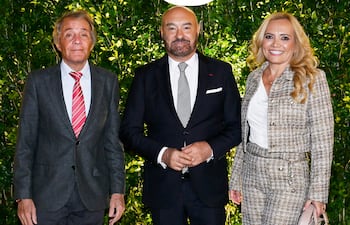Hombre canoso con camisa blanca y corbata roja, hombre con barba en traje oscuro, y mujer rubia en conjunto de tweed sosteniendo un bolso claro.