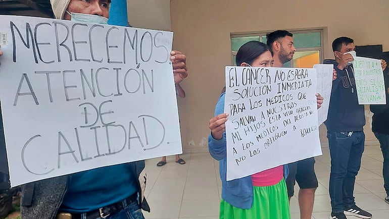 Cuatro padres con mascarillas sostienen carteles en protesta, mientras expresan preocupación por la atención médica en ambiente interior.