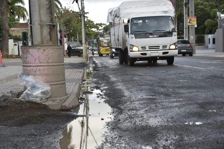 Camión blanco en mal estado de pavimentación, con charcos de agua y árboles al fondo en Avenida España.