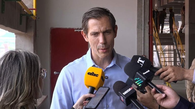 Hombre con camisa clara habla en conferencia; mujer a su lado toma notas, rodeados de micrófonos y ambiente de construcción.