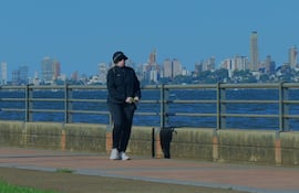 Una mujer vestida con chaqueta negra y gorra camina por el paseo costero en un día soleado y frío.