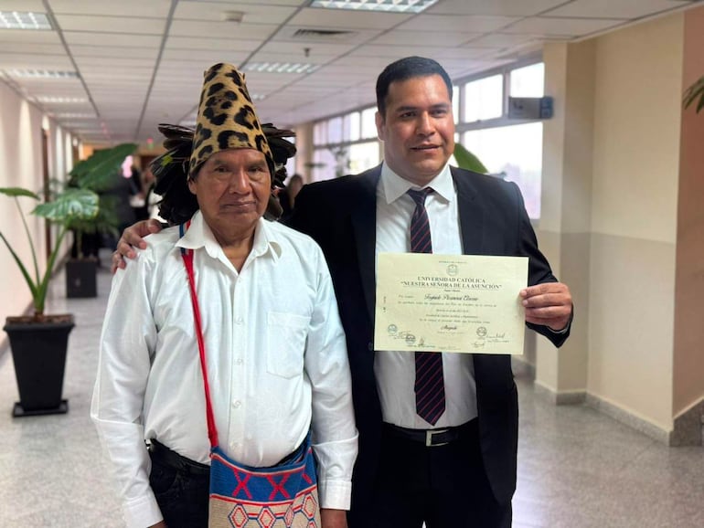 Hombre mayor con atuendo tradicional y mochila artesanal, junto a otro hombre con traje oscuro sosteniendo un certificado en ambiente formal.