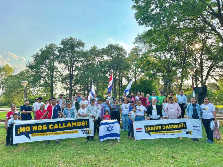 Grupo de personas con camisetas blancas y banderas sostiene pancartas, mostrando expresiones alegres en un parque.