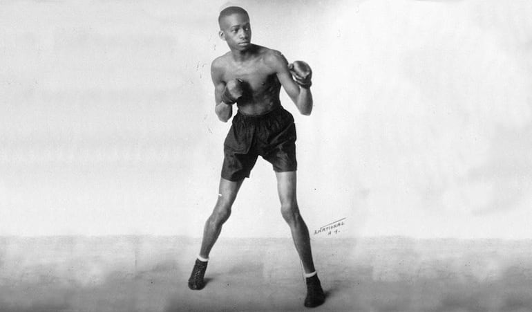 Panamá Al Brown en posición de combate, vistiendo pantalones cortos oscuros y calzado de boxeo, con fondo neutro.