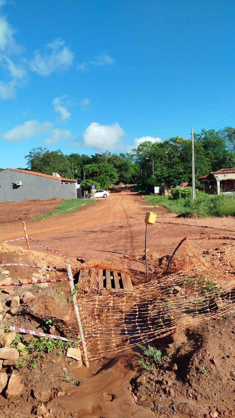 Calle de tierra con materiales de construcción, área de trabajo y casas al fondo. Cielo despejado con algunas nubes.