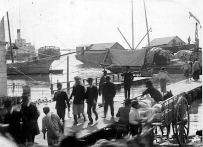Hombres en trajes oscuros de pie en muelle; barcos y carro de carga al fondo, en ambiente de actividad portuaria.