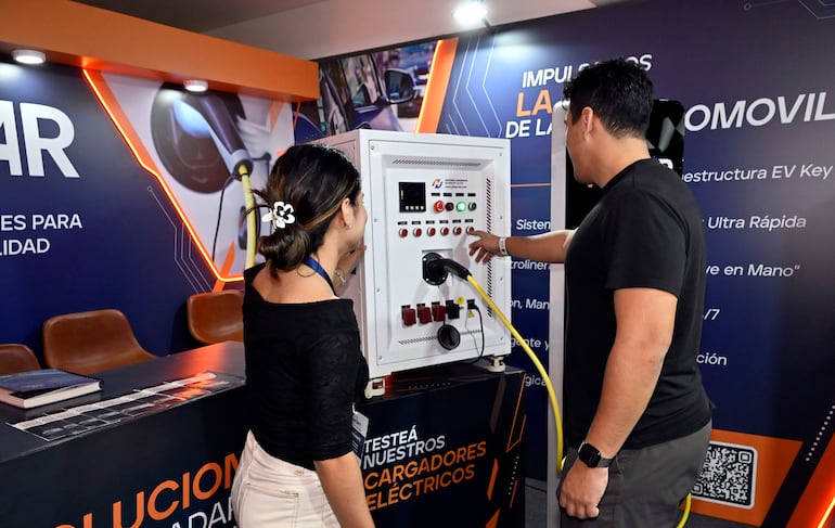 Mujer en blusa negra y hombre en camiseta negra observan un cargador eléctrico en un stand con gráficos de movilidad.