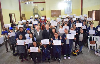 Mujeres sonrientes sosteniendo certificados en alto en salón amarillo, algunas con gorras y chaquetas, en ambiente festivo.