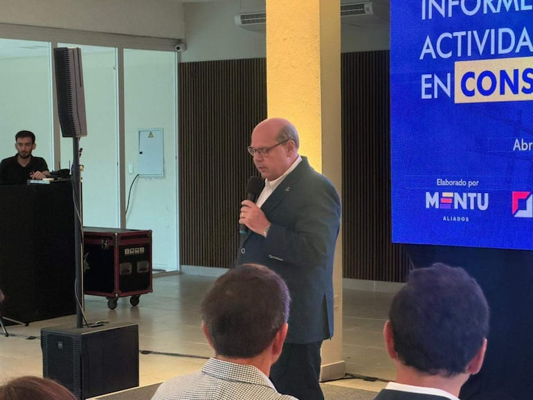 Hombre de mediana edad con gafas habla al micrófono en un evento. Asistentes adultos lo escuchan atentamente en un ambiente formal.