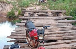 Motocicleta roja estacionada en un puente de madera sobre agua, rodeada de densa vegetación y un entorno natural.