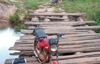 Motocicleta roja estacionada en un puente de madera sobre agua, rodeada de densa vegetación y un entorno natural.
