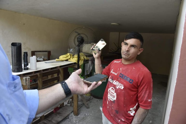 Hombre con camiseta roja de 'Cerro' sostiene un objeto metálico mientras otro usa un teléfono móvil en un taller sencillo.