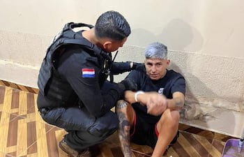 Hombre en chaleco de seguridad agachado cerca de otro hombre canoso sentado, en un ambiente de pared manchada.