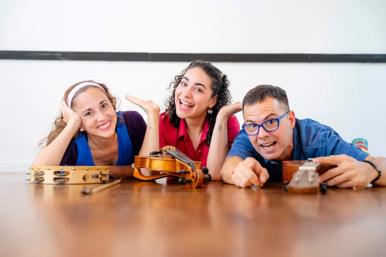 Mujer de blusa rosa sonriente al centro, acompañada de otra mujer en blusa azul y un hombre con gafas, rodeados de instrumentos.