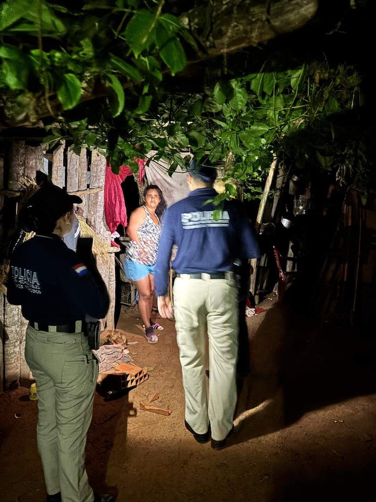 Mujer con cabello rizado y blusa oscura dialoga con dos agentes de policía en un ambiente precario y nocturno.