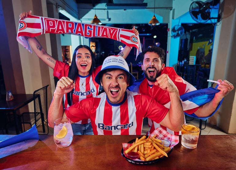 Tres personas en camisetas rojas y blancas levantan los brazos y una bufanda en un bar festivo, con bebidas y papas fritas en la mesa.