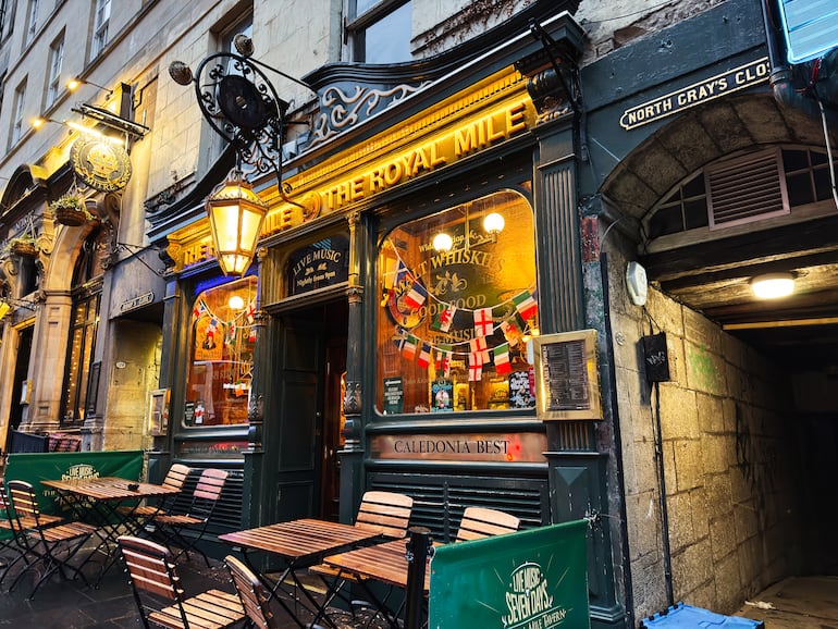 Pub cálidamente iluminado en la Royal Mile de Edimburgo, con mesas al aire libre, banderas, un histórico pasaje de piedra y un ambiente acogedor.