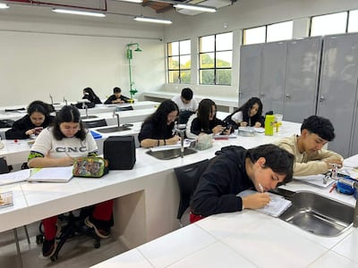 Ocho estudiantes en aula iluminada; una mujer en el centro sostiene una cámara, mientras otros escriben y dibujan en sus cuadernos.