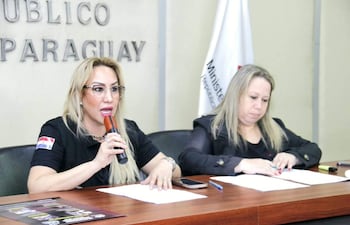 Sandra Ledesma con micrófono y camiseta negra con bandera paraguaya, Karina Cerón sentada, ambas en conferencia formal, ambiente gubernamental.