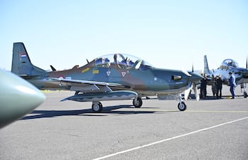 Dos pilotos en la cabina del Super Tucano mientras un grupo de personas en uniforme y civil discute alrededor en un día soleado.