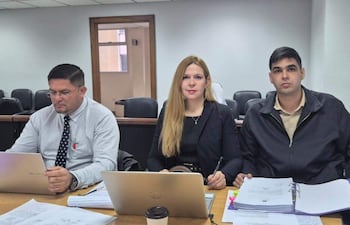 Mujer de cabello largo y rubio en blazer negro observa a la cámara, mientras hombres en camisas gris y chaqueta oscura trabajan en laptops.