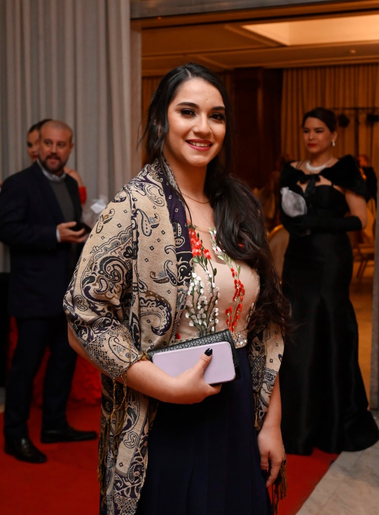 Mujer de pie sonriente con vestido floral y chal oscuro, sosteniendo un bolso en un evento formal con decoración sofisticada.