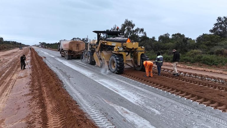 Trabajadores en overol naranja y uniforme en cuclillas, observando el asfalto con maquinaria pesada al fondo en un entorno rural.