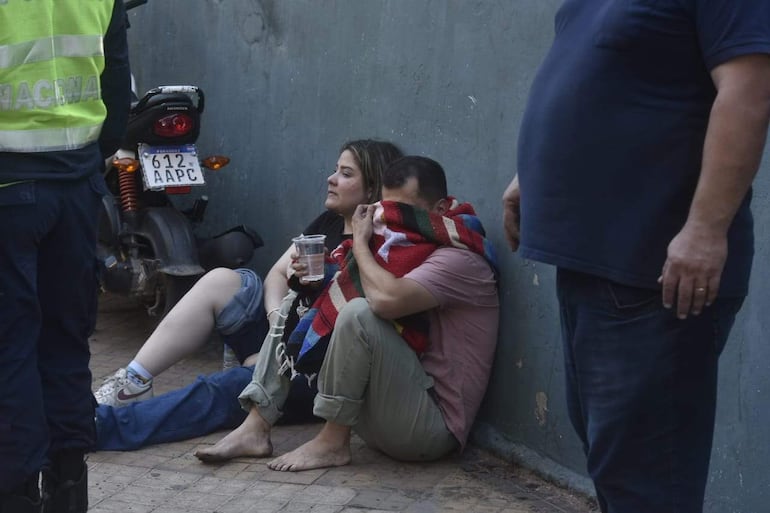 Mujer y hombre sentados en el suelo con expresión de angustia, mientras un oficial con chaleco reflectante les asiste en un entorno urbano.