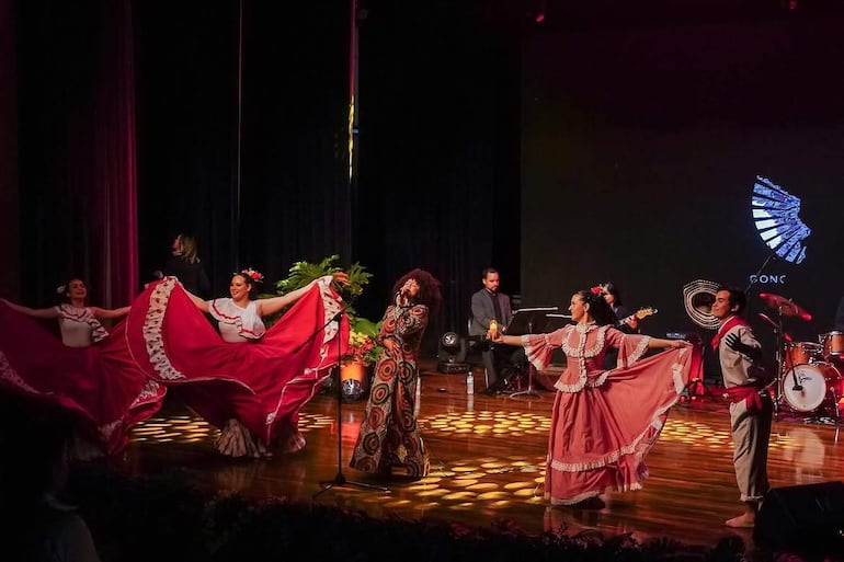 Concha Bernal en vestido colorido rodeada de bailarinas en trajes rojos y beige, con música en vivo en un teatro iluminado.