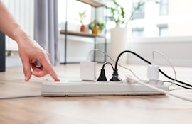 Una mano aprieta el botón de encendido de una zapatilla con cinco enchufes enchufados.