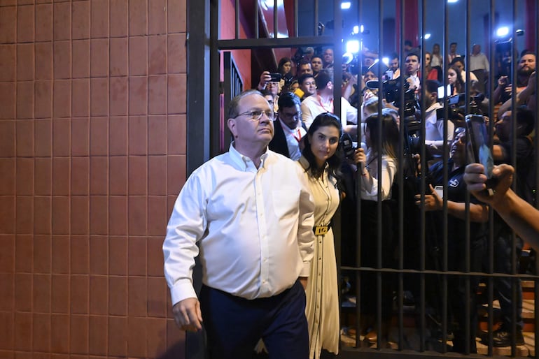 Camilo Pérez con camisa blanca y pantalón oscuro, seguido de una mujer con vestido claro, mientras periodistas capturan su salida.