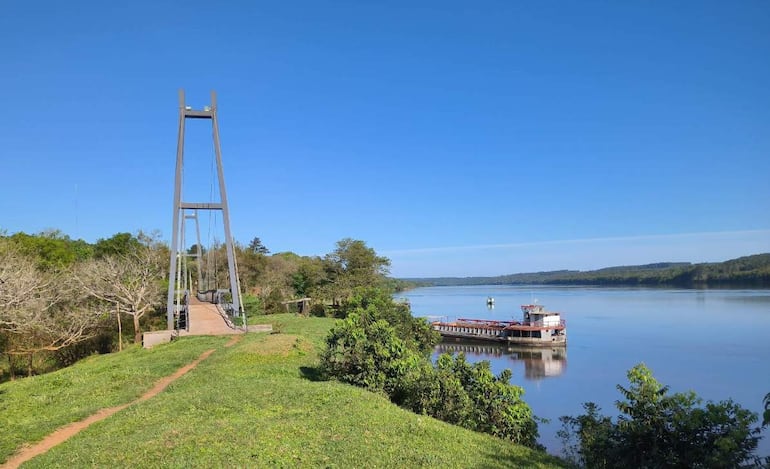 Puente colgante de estructura metálica sobre un paisaje fluvial, rodeado de vegetación densa y un barco de carga atracado.