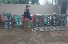 Funcionario de policía de espaldas con uniforme oscuro, rodeado de paquetes de marihuana apilados en un ambiente al aire libre.