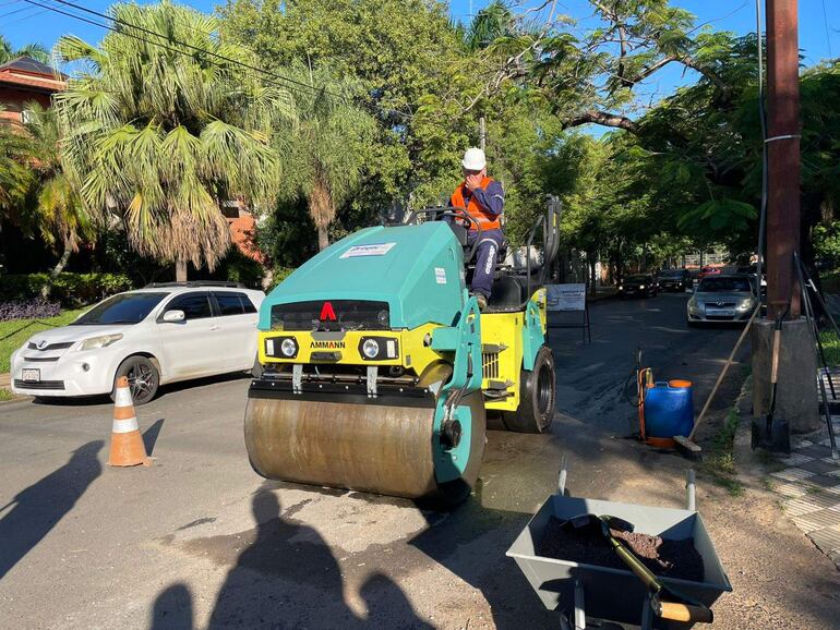Hombre con casco y chaleco de seguridad opera un rodillo Ammann en una calle residencial soleada.