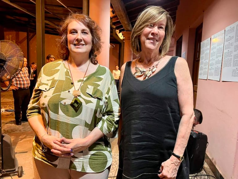 Mujer a la izquierda con blusa verde y cadena, y mujer a la derecha en vestido negro, ambas sonriendo en ambiente cálido.