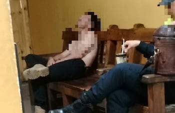 Agente con torso desnudo sentado en un banco junto a otro en atuendo oscuro. Ambiente informal con paredes amarillas.