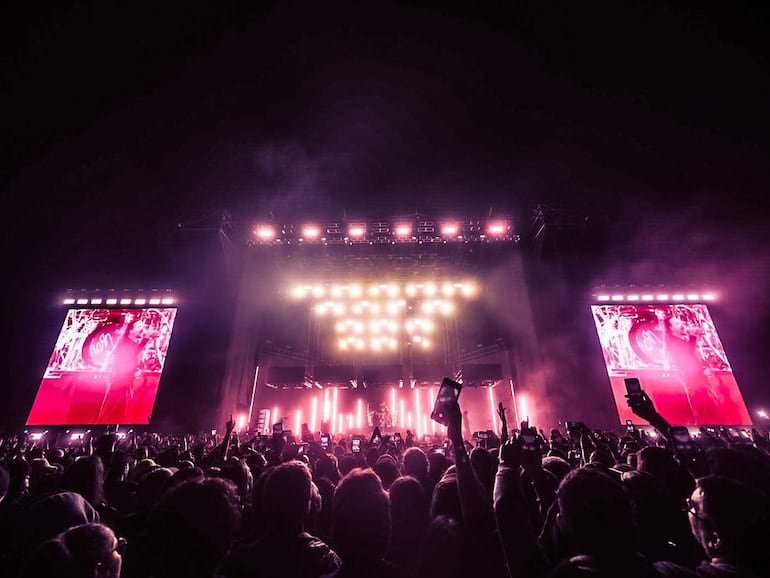 Multitud anónima en concierto de Korn, con teléfonos levantados y luces rosas y blancas iluminando el escenario.