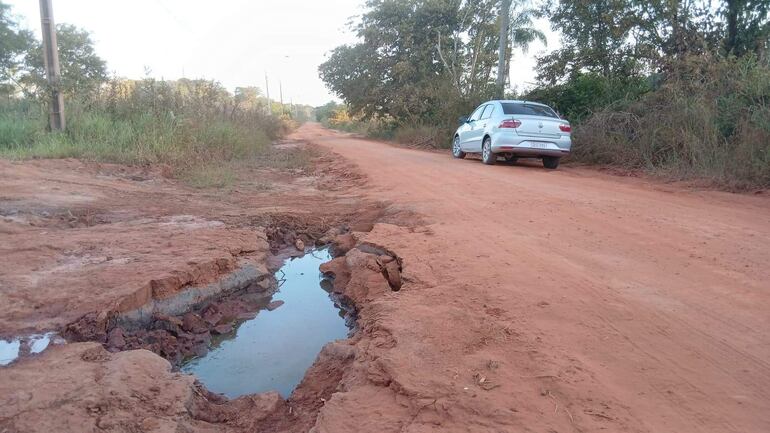 Un camino de tierra con un vehículo blanco estacionado, sin personas, y un bache con agua en un entorno natural.