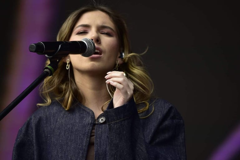 Mavi canta con micrófono, vestida con chaqueta de mezclilla oscura, en un escenario iluminado con luces moradas.