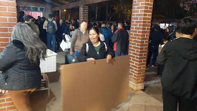Mujer en primer plano sosteniendo cartón, rodeada de otras personas conversando en un ambiente cálido y acogedor.