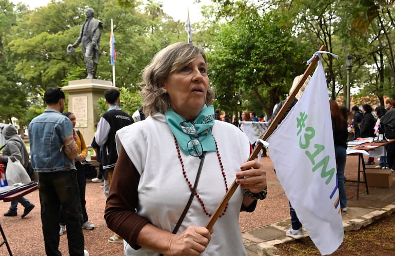 Mujer de cabello canoso y blusa blanca sostiene un palo con bandera, rodeada de personas en un parque durante la marcha.